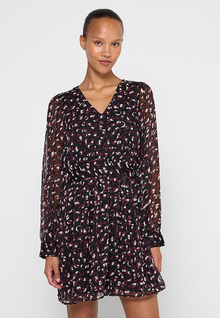 Femme portant une robe noire avec des taches abstraites rouges et blanches, des manches longues transparentes, un col en V et une taille cintrée, se tenant devant un fond blanc uni.