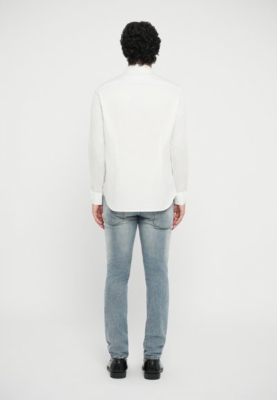 Vit långärmad skjorta med rundad fåll, tillsammans med ljusblå jeans och svarta skor, fångad bakifrån.