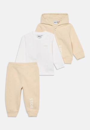 Veste à capuche beige avec fermeture éclair, pantalon de jogging beige avec le texte "BOSS", et chemise blanche à manches longues avec petit logo "BOSS" sur la poitrine.