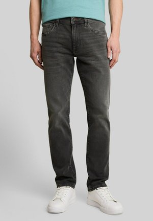 Mann trägt ausgewaschene schwarze Slim-Fit-Jeans, weiße Sneakers und ein hellblaues T-Shirt, steht vor einem neutral grauen Hintergrund.