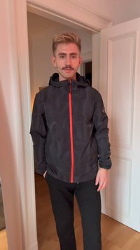 Jovem com cabelo loiro curto e bigode, vestindo uma jaqueta preta com um fecho éclair vermelho, de pé numa porta dentro de uma casa.