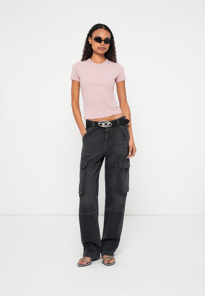 Kvinde med krøllet hår iført sorte solbriller, lavendelfarvet tætsiddende t-shirt, sorte cargobukser med bælte og sorte åbne hæle stående mod hvid baggrund.