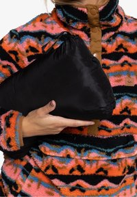 Main tenant une veste en nylon noire pliée devant un pull en polaire à motifs colorés avec des zigzags orange, rose, bleu et noir.