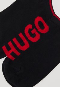 Melni zeķes ar drosmīgu sarkano "HUGO" logotipu priekšā, ar gludu adījuma tekstūru un kontrastējošu sarkanu apdari augšā.