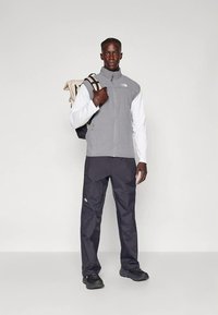 Grå vest med lynlås over en hvid langærmet skjorte, kombineret med sorte bukser. Sorte sneakers og en beige rygsæk med mørke stropper.