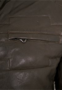 Braune Lederjacke mit einer Reißverschlusstasche auf der Brust, sichtbaren Nähten und einer glatten Textur. Akzente sind ein silberner Reißverschlussanhänger.