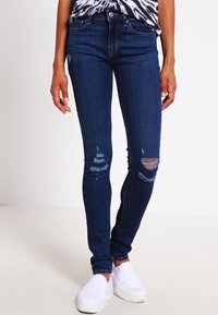 Osoba v tmavomodrých potrhaných slim džínách, černobílém vzorovaném tričku a bílých slip-on teniskách.