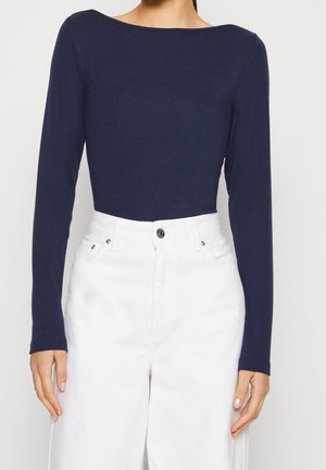 Femme portant un haut ajusté à manches longues bleu marine, rentré dans un pantalon blanc taille haute avec boutons et poches visibles.