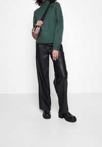 Personne portant un pull à col roulé vert, un pantalon en cuir noir à jambes larges, des bottes épaisses noires et un sac bandoulière imprimé léopard.