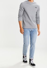 Harmaa huppari, jossa on pyöreä kaula-aukko ja musta logopatchi, yhdistettynä vaaleansinisiin farkkuihin ja mustiin korkeavartisiin tennareihin, jossa on valkoiset nauhat.