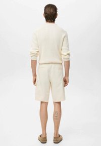 Pull en tricot texturé crème associé à un short beige clair avec des poches. Sandales marron clair, tatouages visibles sur la jambe droite.