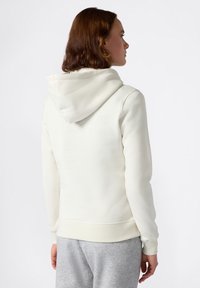 Jonge vrouw met kort bruin haar, gekleed in een eenvoudige witte hoodie en lichtgrijze trainingsbroek, gezien van achteren tegen een witte achtergrond.
