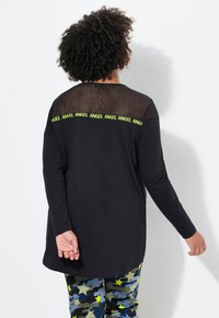 Zwarte langslijfshirt met een mesh rugaccent en een herhalende "ANGEL" grafiek in felgroen. Gecombineerd met camouflageprintbroek.