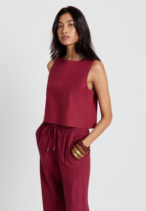 Donna con capelli lunghi e scuri che indossa un top senza maniche color bordeaux e pantaloni coordinati con tasche laterali e braccialetti di legno al polso.