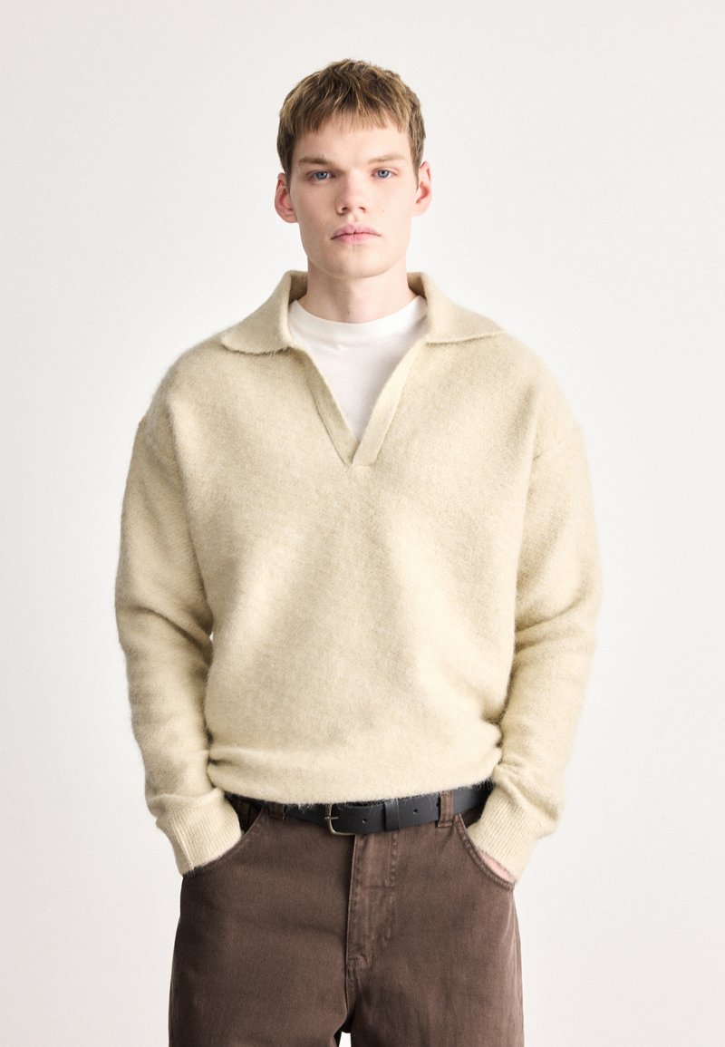 Béžový pletený sveter s výstrihom do V a plochým golierom, vyrobený z mäkkej, textúrovanej látky. Kombinovaný s tmavohnedými nohavicami a čiernym opaskom.