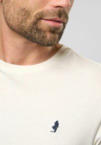 Světle béžové bavlněné tričko s kulatým výstřihem a malou tmavě modrou vyšívanou postavičkou na hrudi. Hladká textura, ležérní design.