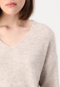 Maglione beige a costine con scollatura a V e vestibilità rilassata, che mostra una morbida texture e sottili variazioni di colore. Confortevole e leggero.