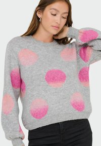 Jeune femme portant un pull gris avec de grands pois roses en dégradé, posant avec une main derrière le cou et les yeux fermés.