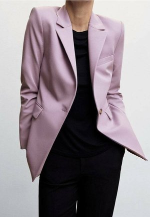 Personne portant un blazer rose clair sur une chemise noire et un pantalon noir sur un fond blanc uni.