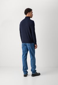 Ναυτικό πουλόβερ με ψηλό γιακά, συνδυασμένο με μπλε τζιν. Το σύνολο διαθέτει χαλαρή εφαρμογή και λεία υφή, συμπληρωμένο από μαύρα παπούτσια.