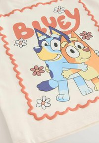 T-shirt bianca con una stampa colorata di due cani dei cartoni animati che si abbracciano, circondati da fiori e un bordo scallop rosa con il testo "BLUEY".