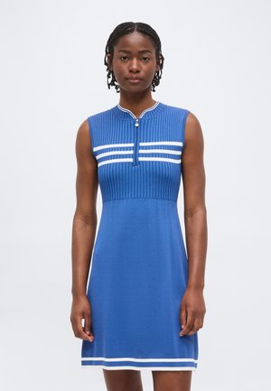 Kobieta z warkoczami, ubrana w niebieską dzianinową sukienkę bez rękawów z białymi paskami i zamkiem błyskawicznym z przodu, stojąca na jednolitym tle.