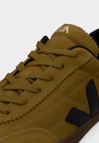 Καφέ σουέτ αθλητικό παπούτσι με υφή στην επιφάνεια, μαύρο λογότυπο και στρογγυλά κορδόνια. Ραφές στις πλευρές και λαστιχένια σόλα.