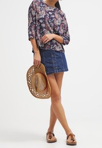 Lös passform i marinblå och paisleymönstrad topp, jeansminikjol med framknappar, halmstuv och bruna sandaler, vilket skapar en avslappnad look.