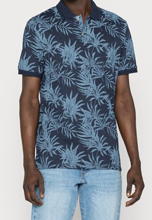 Homme portant un polo bleu marine avec un motif de feuilles tropicales bleu clair et un jean en denim bleu clair, debout avec les bras détendus.