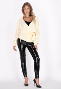 Krämfärgad omslagströja med pärldetaljer på ärmarna, kombinerad med blanka svarta faux läderleggings och svarta högklackade skor.