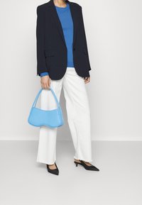 Persona que lleva un blazer negro, un suéter azul, jeans blancos de pierna ancha, tacones negros de punta y sostiene un bolso azul claro con un diseño curvado.