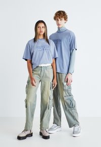 Lyseblå oversized t-skjorter med tekstgrafikk, kombinert med baggy, falmede grønne cargo-bukser med flere lommer. Fottøyet inkluderer kraftige støvler.