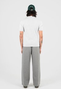 Persona con lunghi capelli ricci che indossa un cappellino verde, una maglietta grigia chiara a maniche corte, pantaloni grigi larghi e tatuaggi visibili su entrambi i bracci.