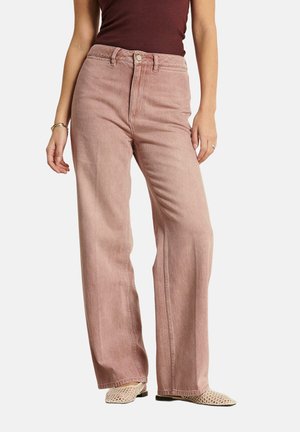 Femme portant un jean rose poussiéreux taille haute à jambes larges, un haut sans manches bordeaux, et des chaussures beige tissées à enfiler, debout les mains détendues.