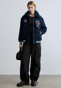 Tamsiai mėlynas universiteto stiliaus džinsinis švarkas su pūstu apykakle, baltomis juostelėmis rankogaliuose ir siuvinėtais elementais. Derinamas su juodomis krovinių kelnėmis ir platforminiais batais.