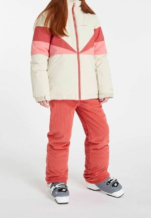 Veste de ski crème et rose avec un design géométrique, associée à un pantalon en velours côtelé rouge. La tenue comprend des chaussures de ski grises avec des accents noirs.