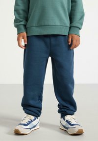 Pantaloni della tuta blu scuro realizzati in tessuto morbido, con vita elasticizzata, orli a costine e una vestibilità comoda. Abbinati a sneakers blu e bianche.