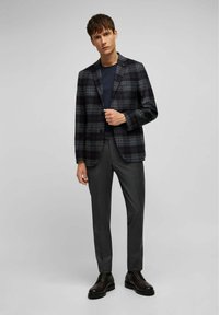 Mannenblazer in donkere ruit met groen, bordeaux en navy. Gecombineerd met donkere broek en zwarte schoenen. Soepele stof met een gestructureerde pasvorm.