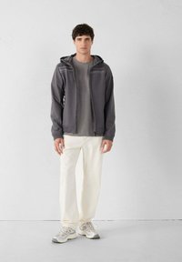 Grijze zip-up jas met capuchon, gecombineerd met een grijs T-shirt, witte broek en lichtgrijze sneakers. Gladde stof, nauwsluitend design, neutrale kleuren.
