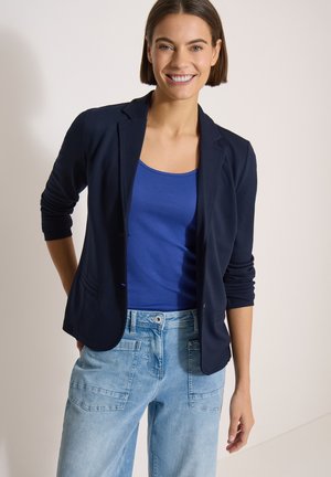 Femme souriante, portant un blazer noir sur un débardeur bleu et un jean bleu clair avec de grandes poches avant, debout devant un fond uni.