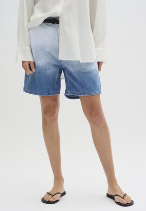 Personne portant un short en jean ombré bleu, une chemise blanche à manches longues et des tongs noires, debout devant un fond blanc.