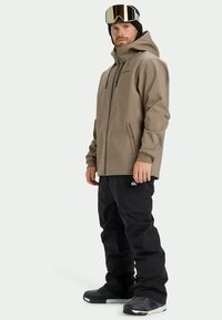 Beige wasserdichte Jacke mit Kapuze, Frontreißverschluss und Seitentaschen, kombiniert mit schwarzen Schneehosen und dunklem Schuhwerk, geeignet für Wintersport.