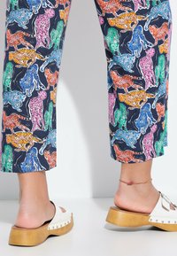 Farverige dyremønstrede bukser på en marineblå baggrund; fod i en hvid sandale med glid og en træplatform med sølvnitter.