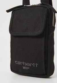 Pochette nera in nylon con pattina, chiusura con zip e logo. Forma rettangolare, superficie testurizzata e tracolla regolabile per il trasporto.