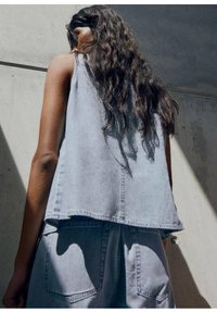 Top sin mangas de denim gris claro con un ajuste relajado, que presenta un dobladillo asimétrico y sutilezas en los detalles de la costura. La tela texturizada capta la luz.