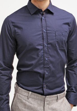 Hombre que lleva una camisa azul oscuro de manga larga con bolsillo en el pecho y pantalones gris claro.