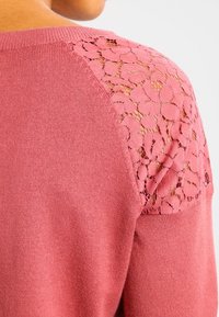 Suéter de punto rosa con detalles de encaje en los hombros, de textura suave y cuerpo sólido. El suéter tiene un escote redondo.