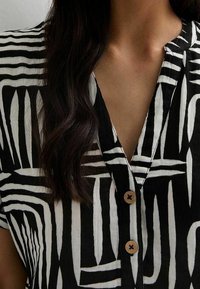 Blusa a maniche lunghe in bianco e nero con scollo a V, caratterizzata da una combinazione di righe orizzontali e verticali. Presenta due bottoni in legno all'annodatura del collo.