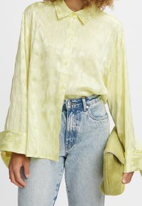 Camicia di seta verde chiaro con una texture lucida, chiusura a bottoni davanti e ampie maniche, abbinata a jeans in denim blu strappati e una pochette coordinata.