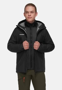 Giacca nera isolante con interno grigio, dotata di cappuccio, chiusura con zip e logo "Mammut" sul lato sinistro del petto, indossata da un modello.
